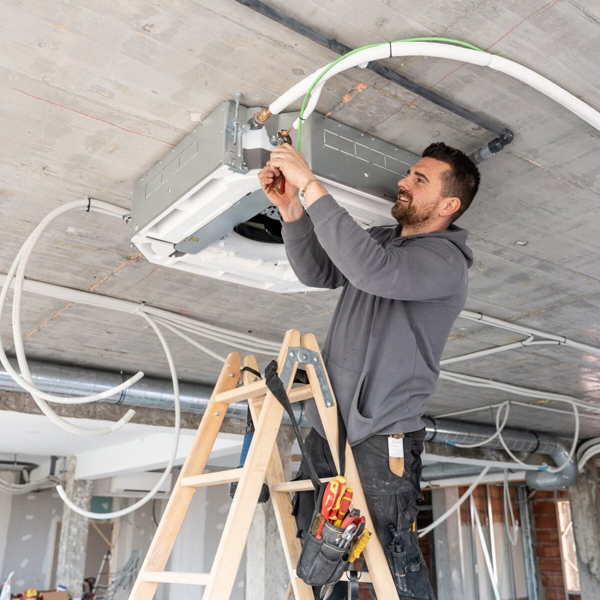 Mannelijke verwarmings- en ventilatietechnicus op een ladder die een nieuwe plafond-airconditioningunit installeert op een bouwplaats, terwijl hij met gereedschap werkt